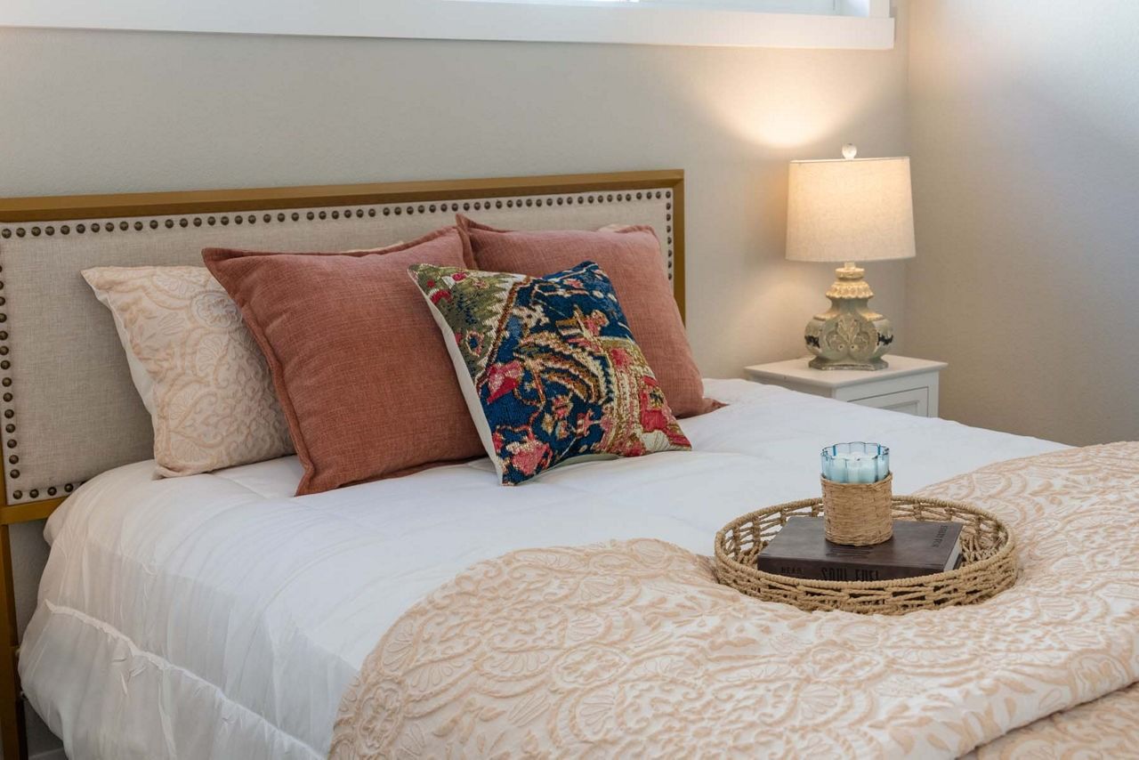 Bright bedroom with a white bed, pink and floral pillows, and a bedside lamp on a white nightstand