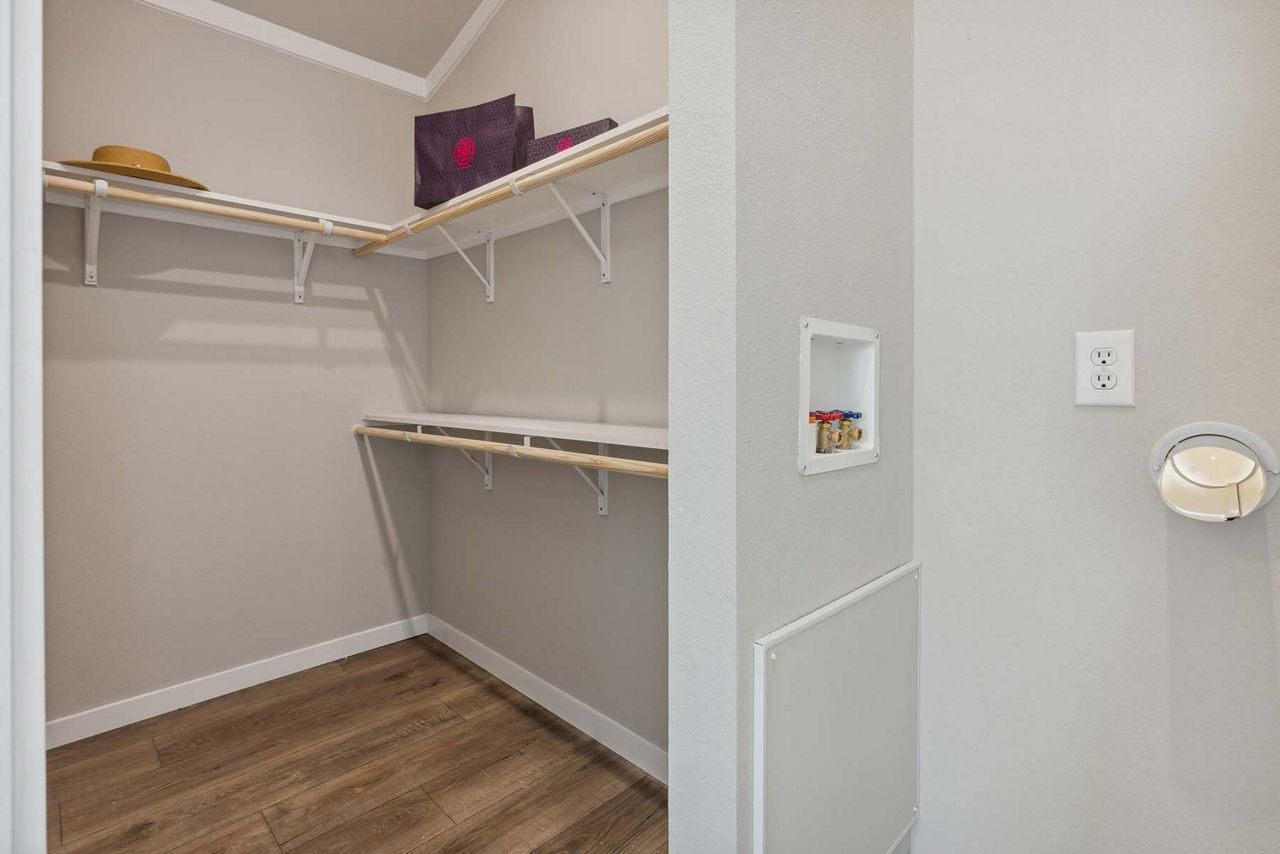 Walk-in closet with white shelves and wood flooring, viewed from an adjacent room with light switch and thermostat.