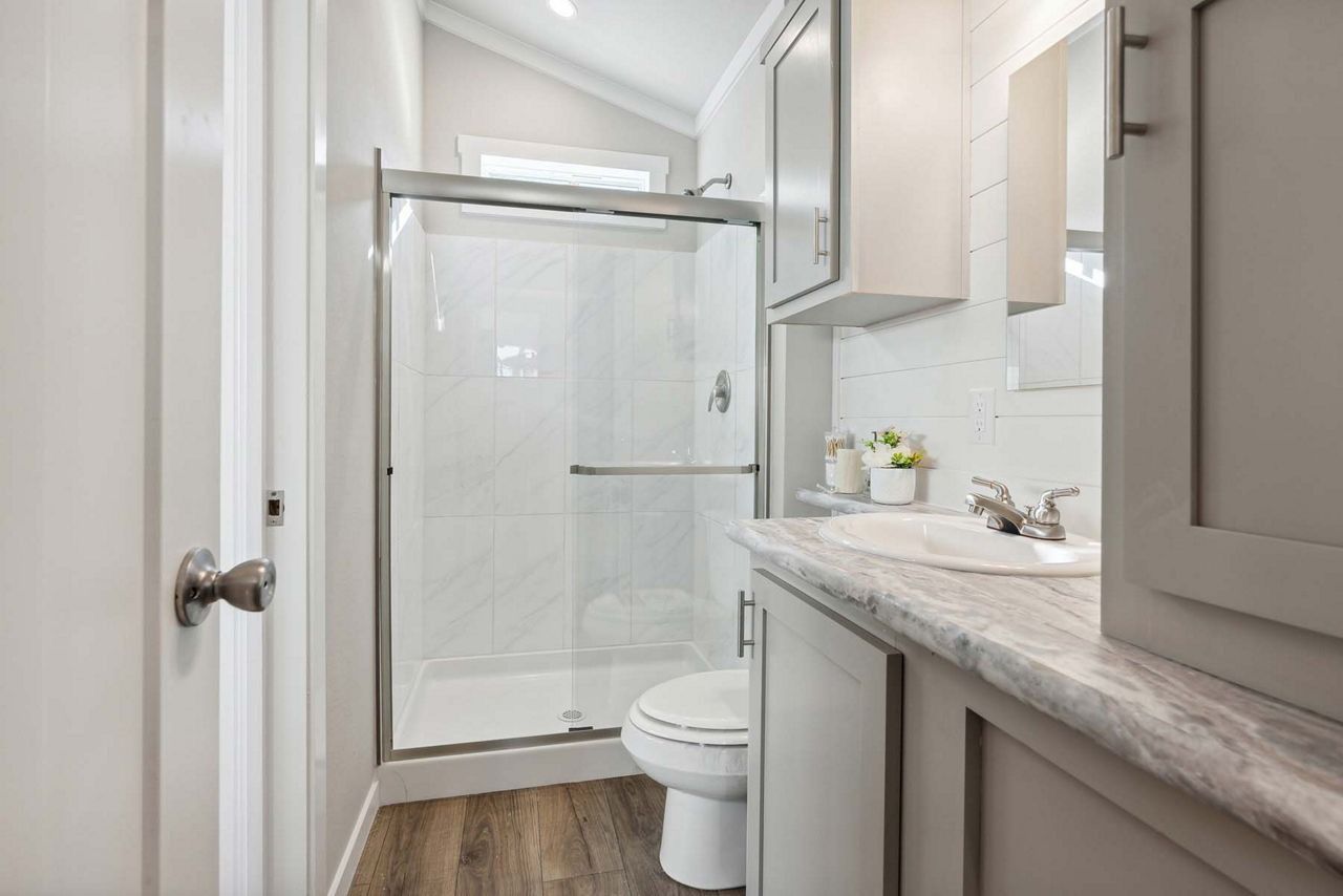 Bright narrow bathroom with white tile, glass shower, toilet, and marble vanity.