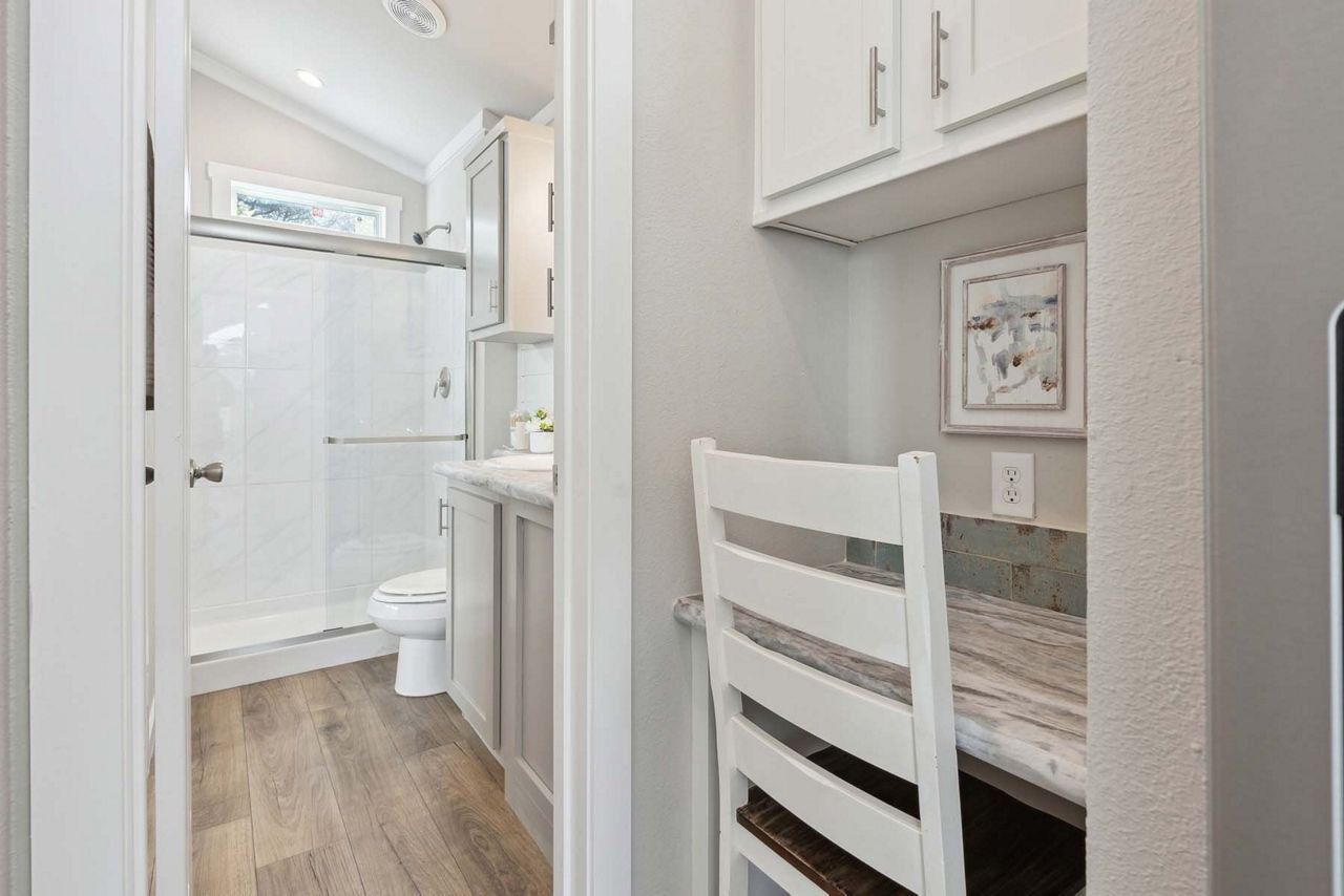 Narrow bathroom with white vanity, chair, shower, and light wood flooring.
