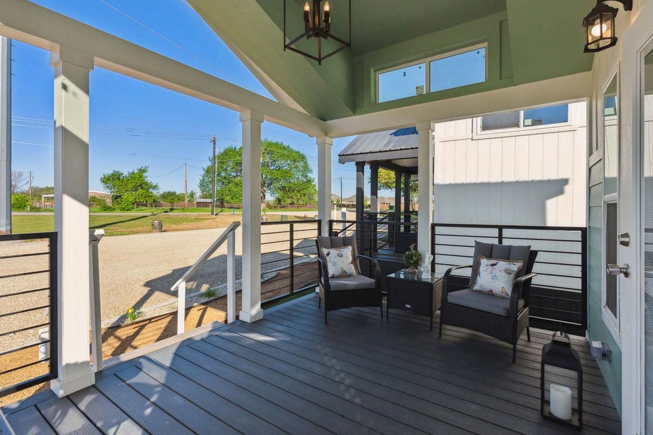 Covered porch with dark wicker chairs, lantern lights, and open views of a sunny yard and street.