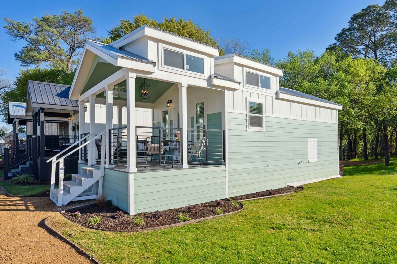 Mint-green tiny house with covered porch and stairs in a grassy yard among trees