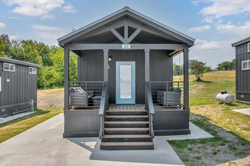Small gray porch cabin with front steps, white trim, and blue door on a grassy lot