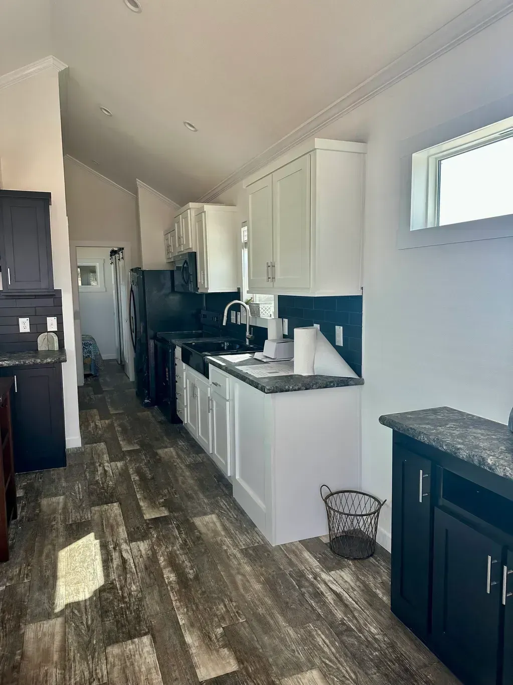 A narrow kitchen with white cabinets, dark countertops, and wood-patterned flooring, featuring an open doorway ahead.