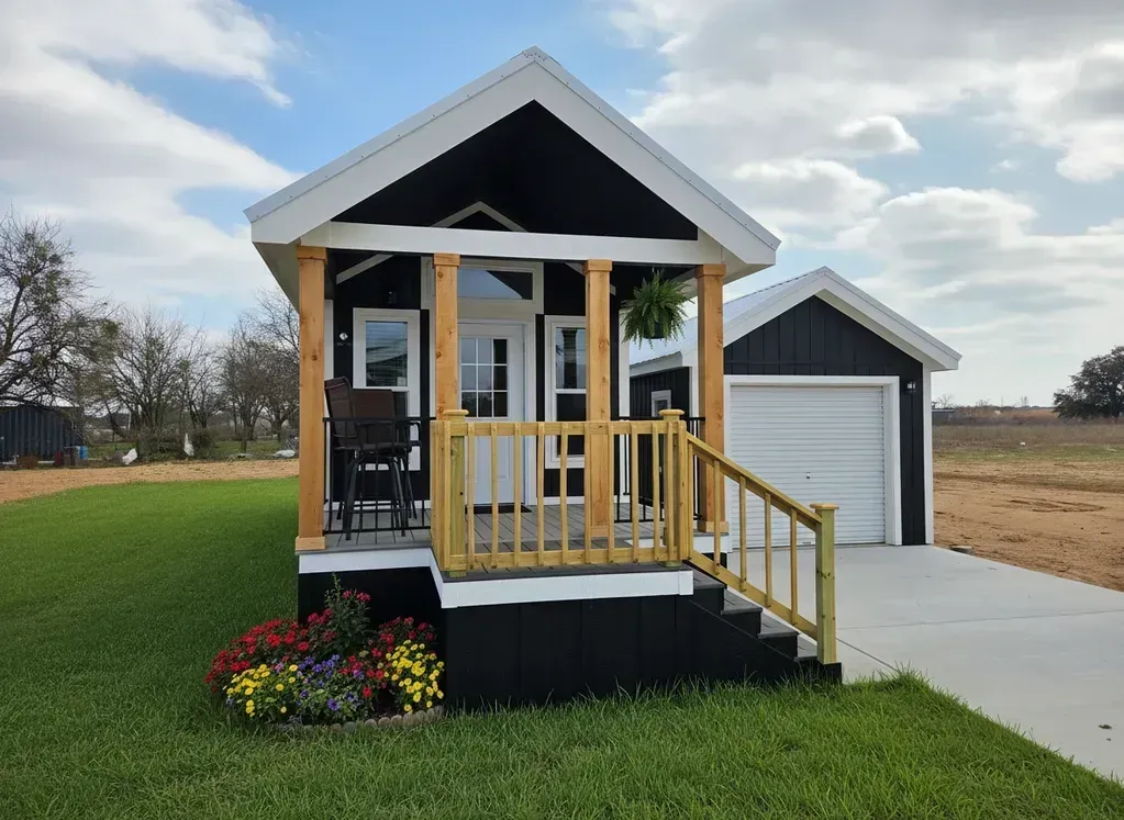 Small white tiny house with yellow porch railing beside a detached garage on a grassy lot