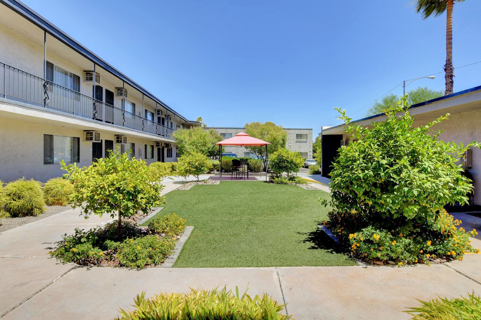 Apartment courtyard with green lawn, shrubs, benches, and a red umbrella under a sunny blue sky