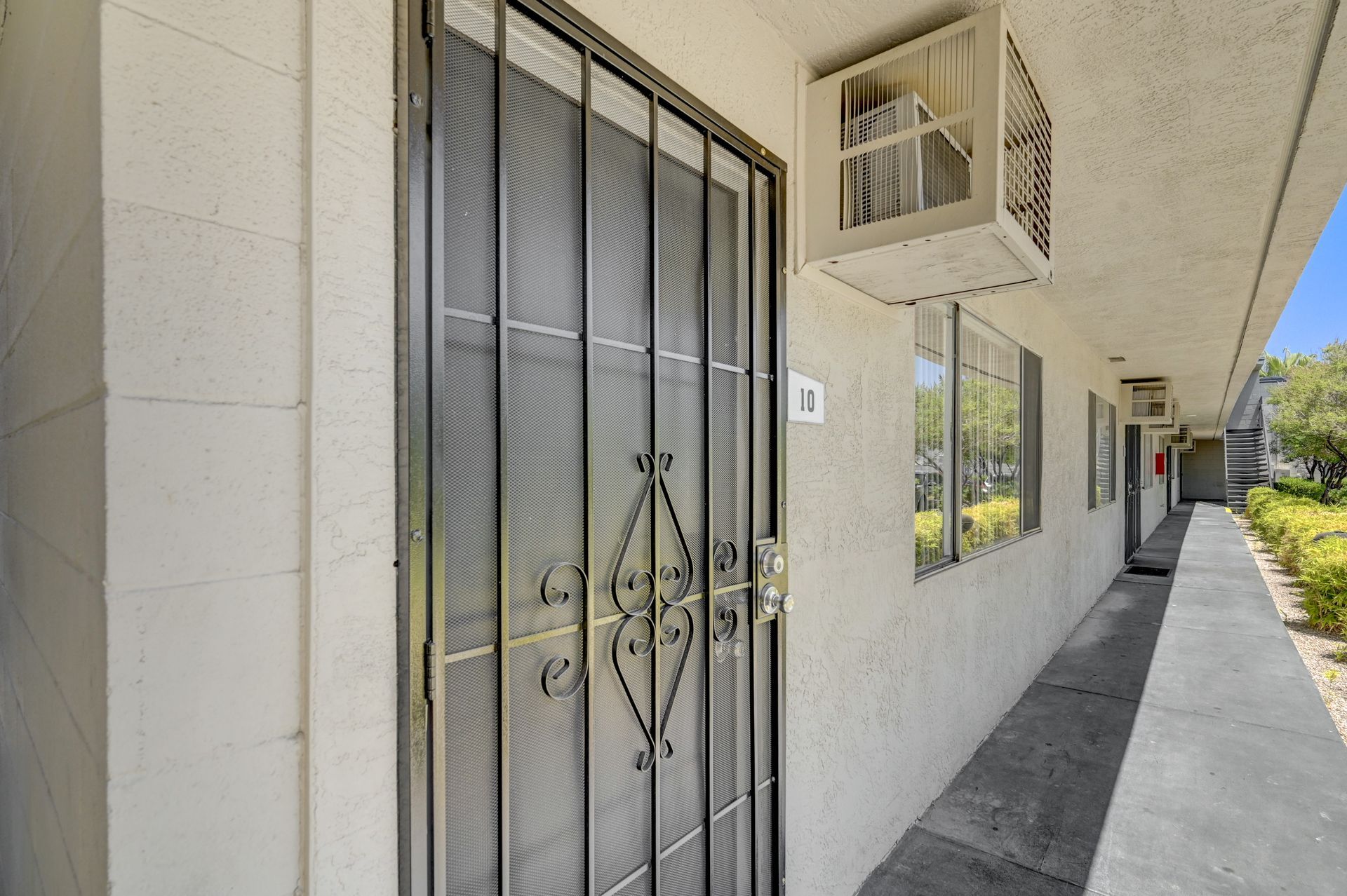 Apartment corridor with gray metal security door, barred window, and white stucco walls under bright sunlight