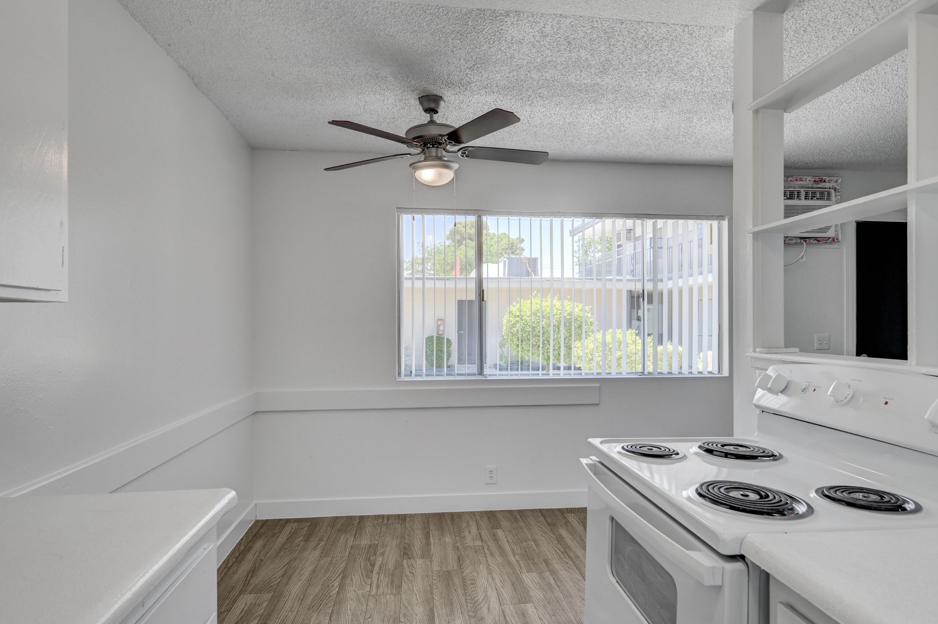 Bright empty kitchen with white appliances, ceiling fan, and large window with vertical blinds.