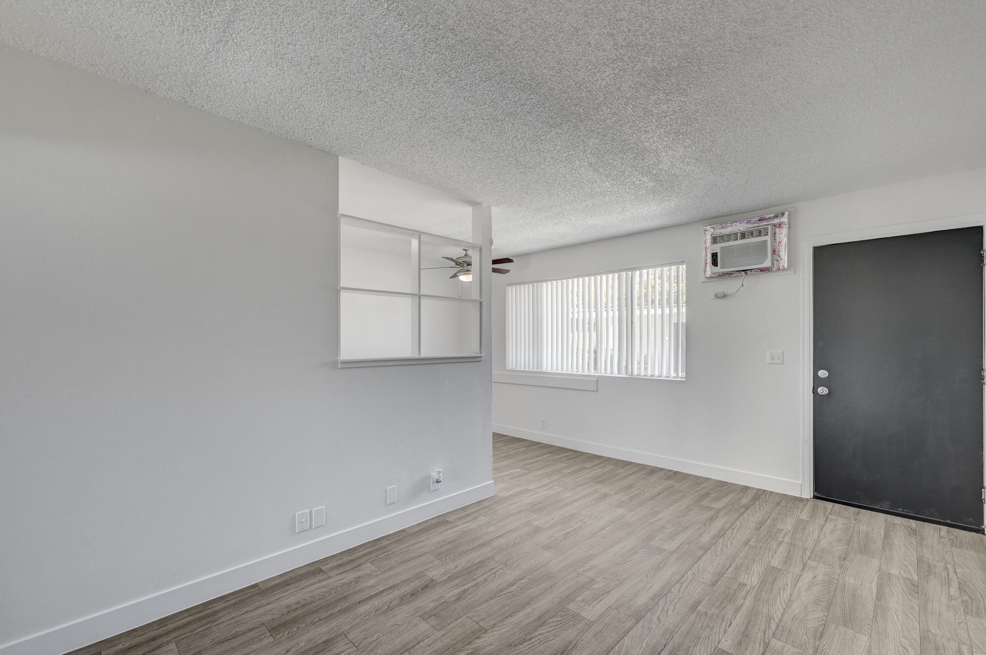 Empty light gray room with wood-look floor, white window blinds, wall cabinets, and a dark door