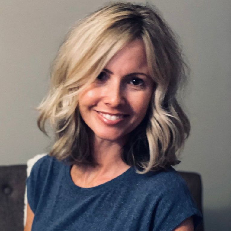 Woman with blonde hair smiles at the camera, wearing a white top. Indoors, lit by warm light.