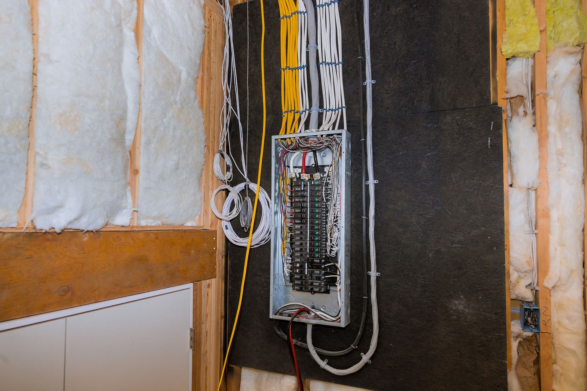 Electrical panel with exposed wiring mounted on a partially unfinished wall inside a building.