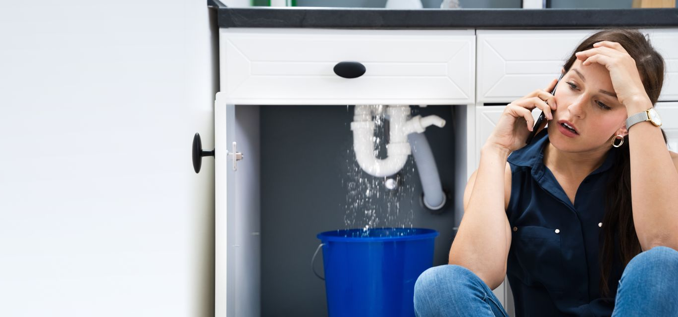 A woman sitting on the floor next to a leaking kitchen sink calling an emergency plumber