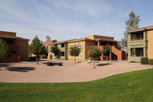 A tan and olive apartment complex featuring an open courtyard with gravel, small trees, and a grassy foreground.