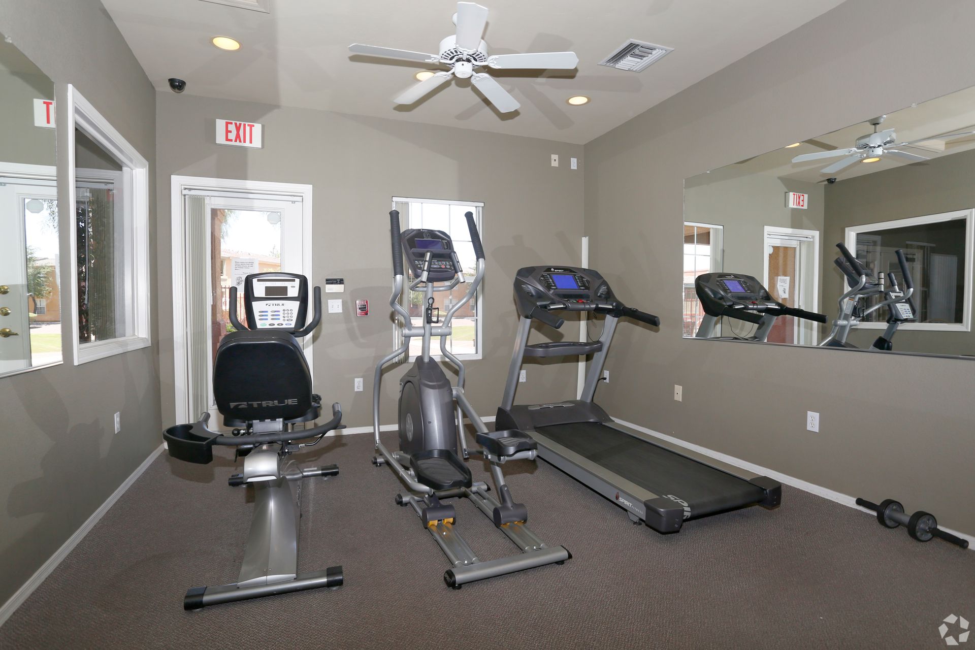 A small gym with an exercise bike, an elliptical, and a treadmill, featuring tan walls, carpet, and a large mirror.