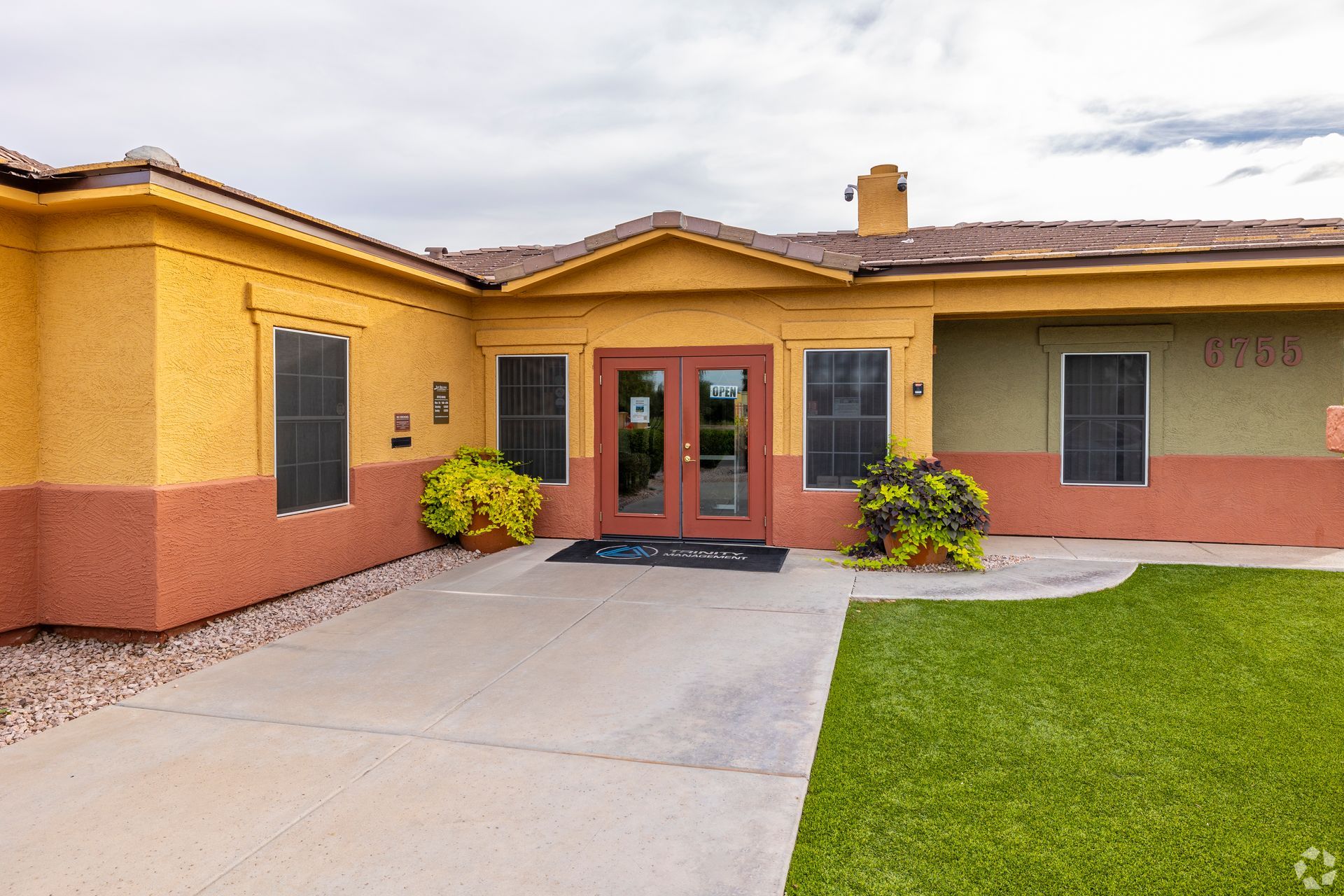 A single-story building with yellow and terracotta stucco walls, double glass entry doors, and a green lawn.