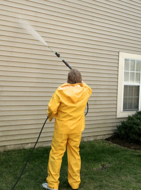 Person in yellow rain suit pressure-washing a house exterior and window.