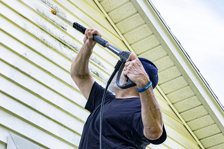Man in a blue cap using a pressure washer on a house’s white siding