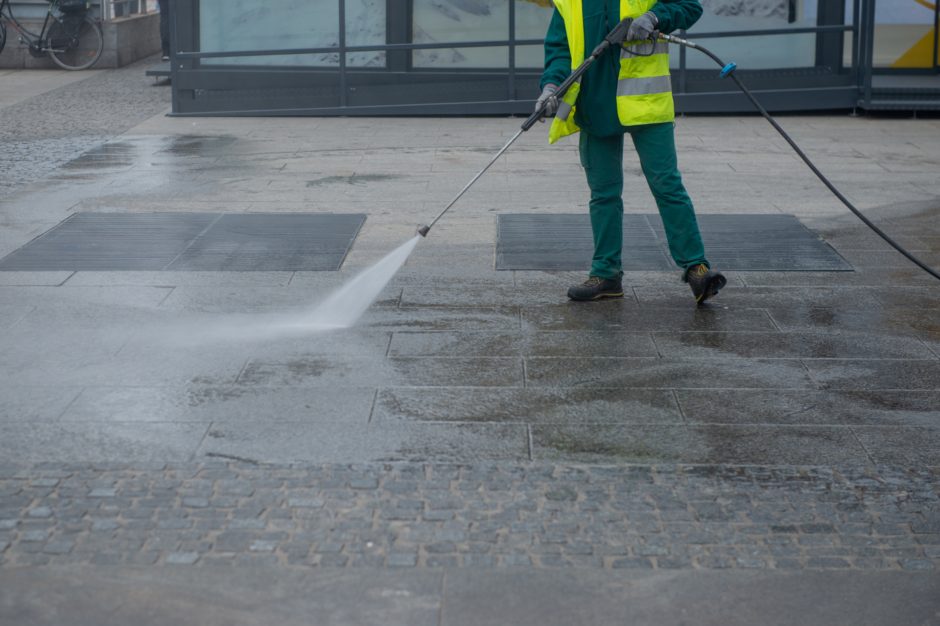 Worker in yellow safety vest pressure-washing a wet sidewalk on a rainy street.