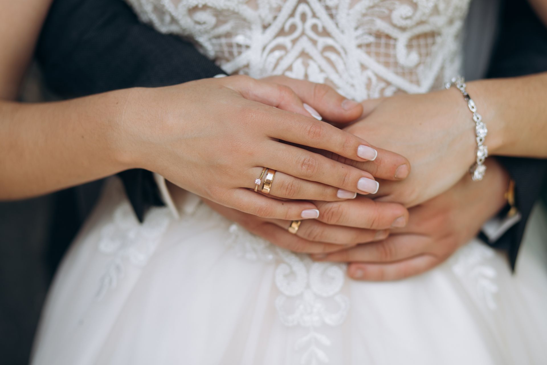 Two people holding hands, showing wedding rings and a white lace dress.