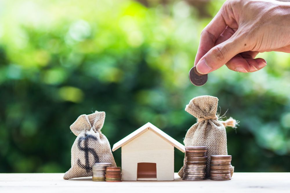 A person is putting a coin into a bag of money next to a model house and stacks of coins.
