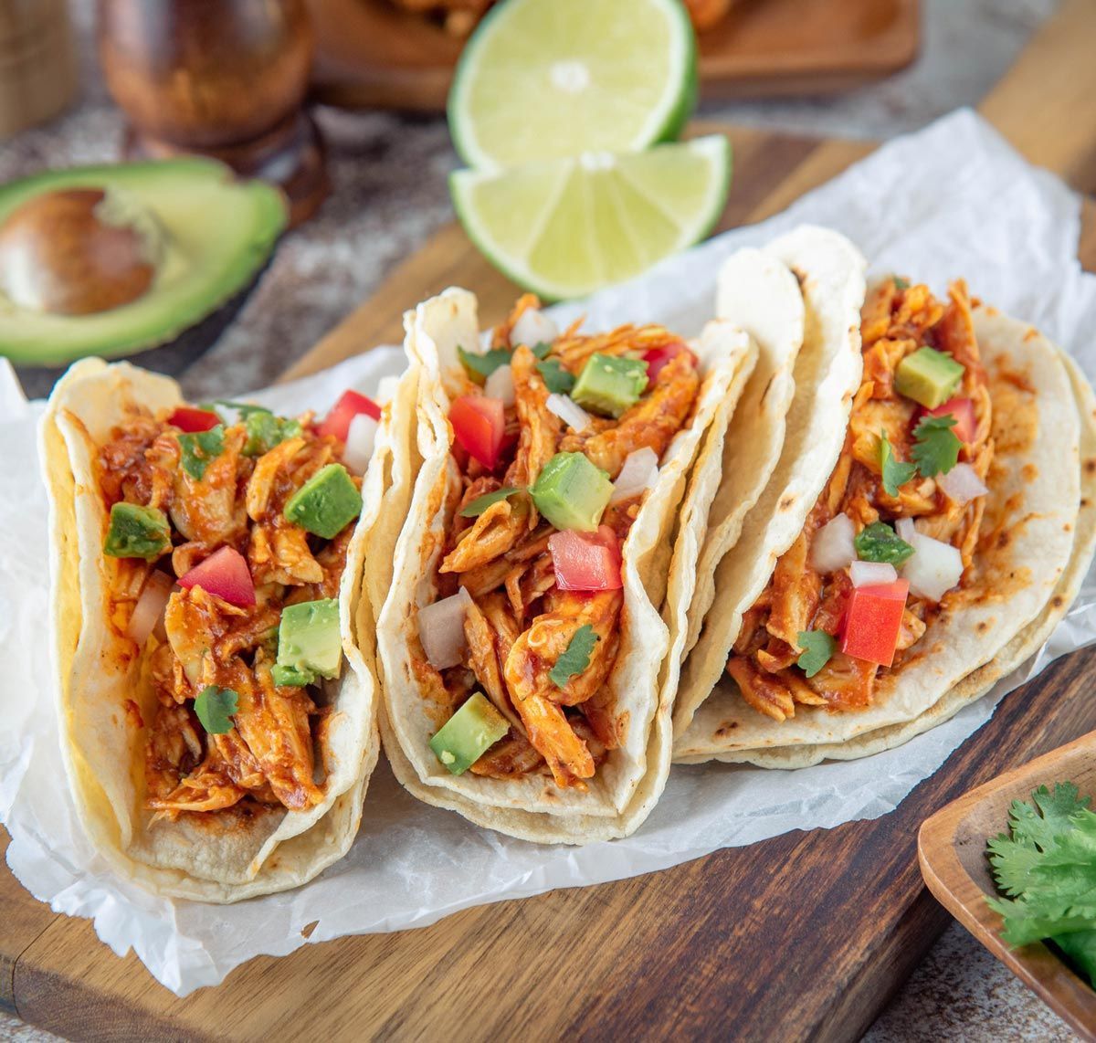 A row of tacos with chicken, avocado, tomatoes and onions on a wooden cutting board.