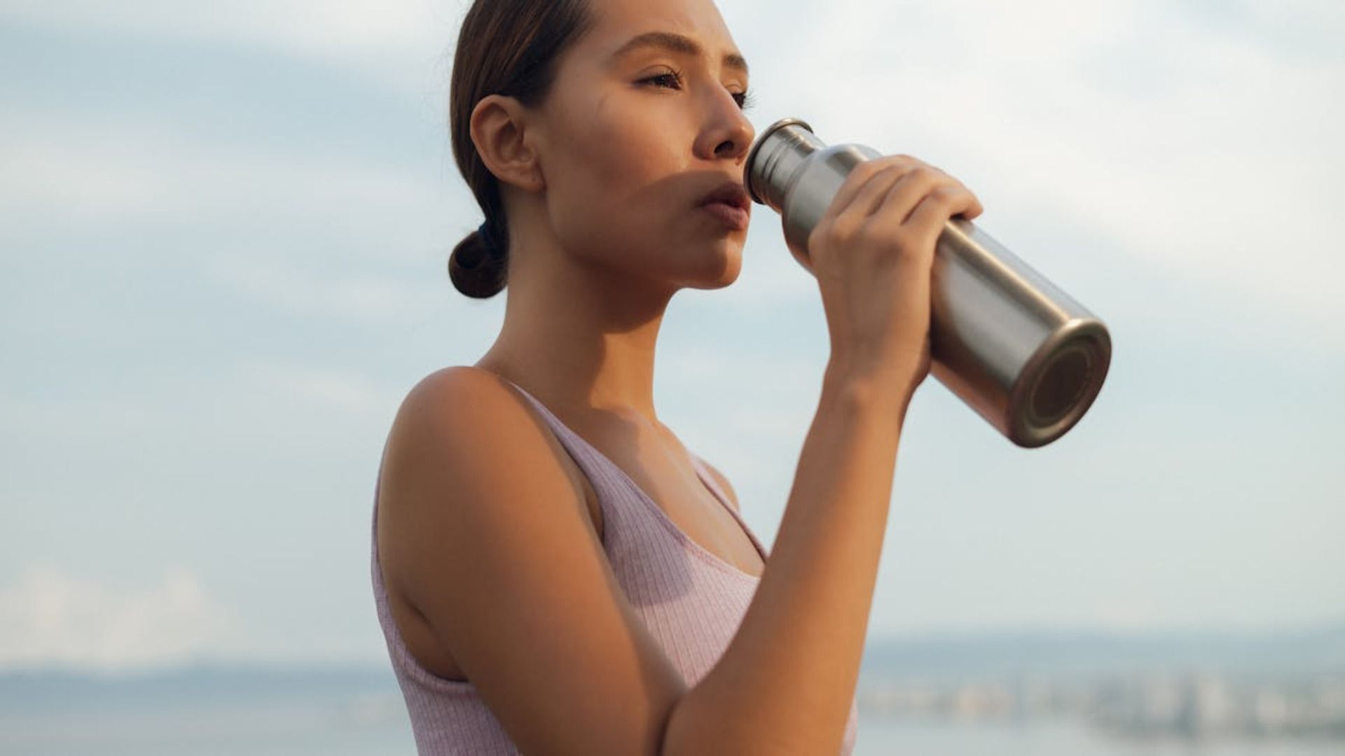Mujer bebiendo de una botella de agua de acero inoxidable al aire libre, agua en el fondo.