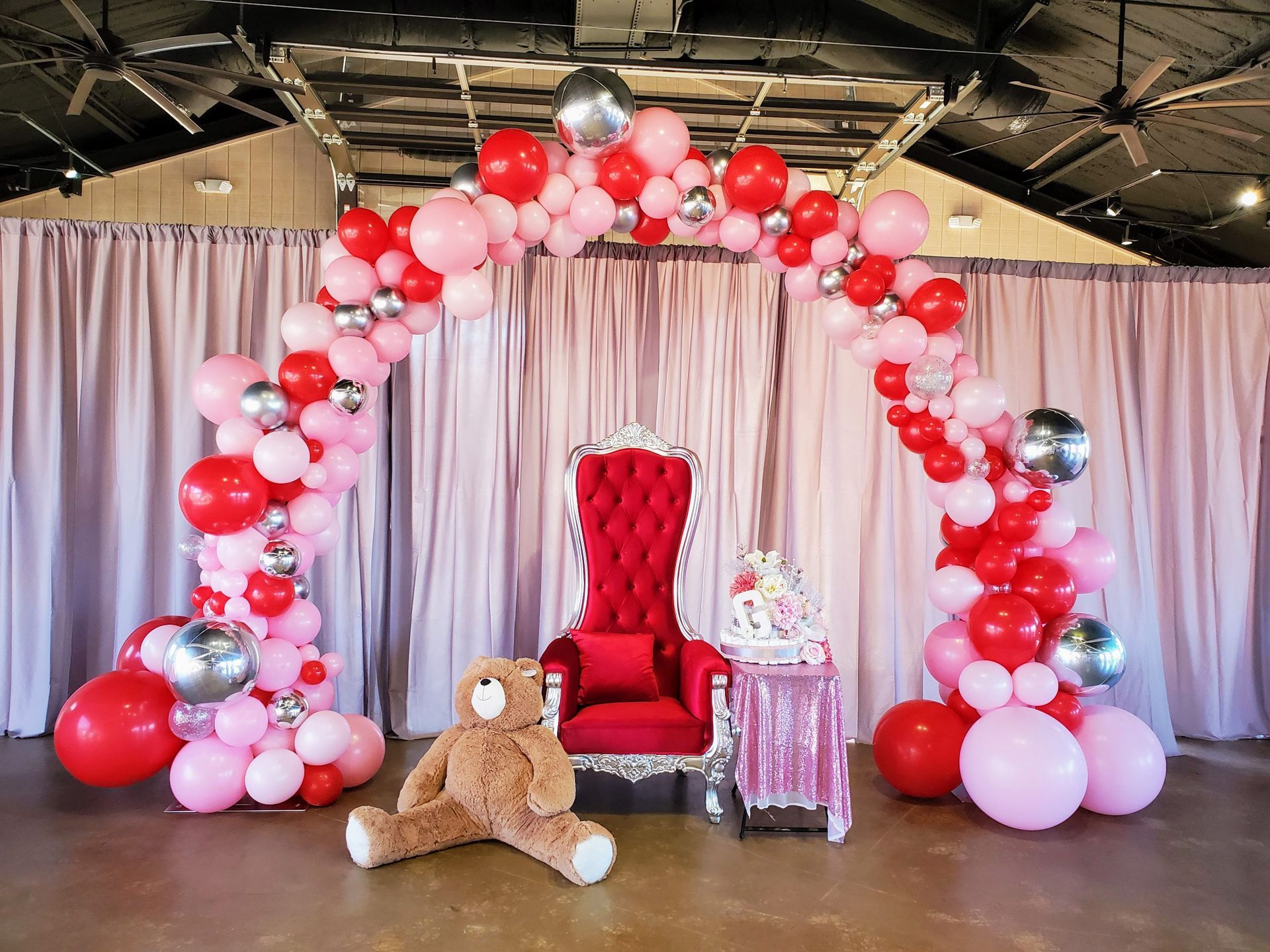 A teddy bear is sitting in a chair in front of a balloon arch.
