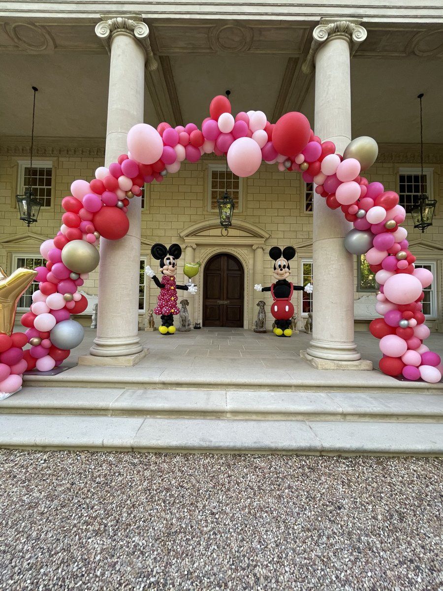 Mickey mouse and minnie mouse are standing under a balloon arch in front of a building.