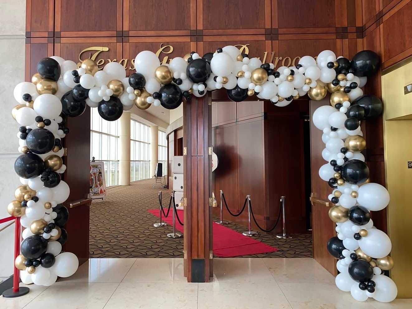 A large arch made of black , white and gold balloons in a room with a red carpet.