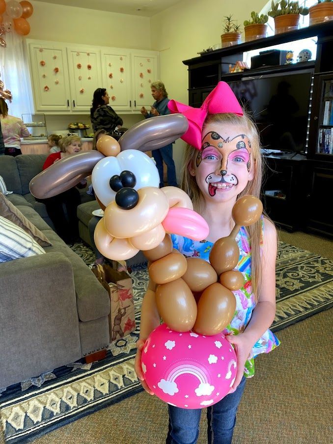 A little girl with balloons painted on her face is holding a balloon in the shape of a dog.
