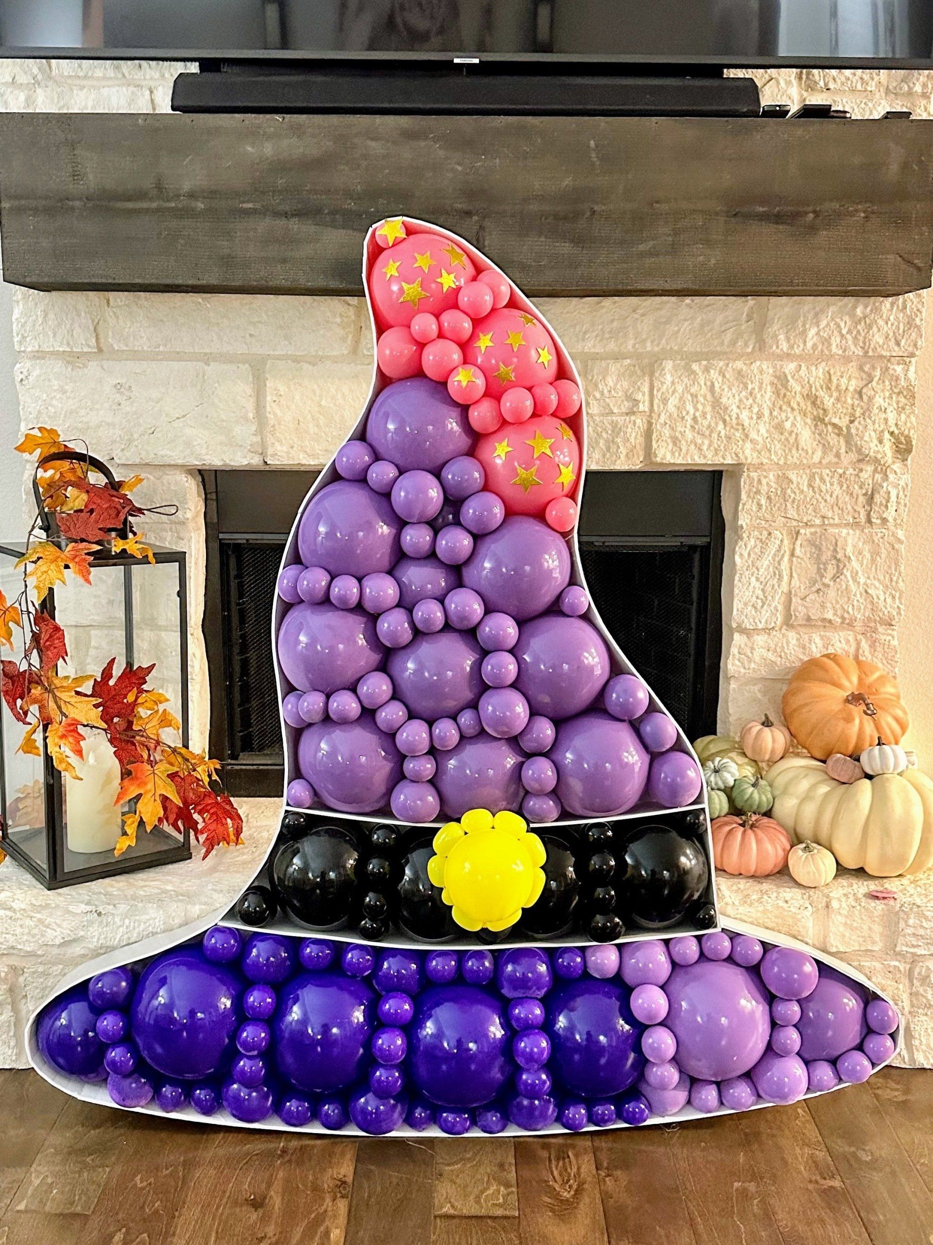 Balloon mosaic of a witch's hat in pink, lilac and black balloons in front of a fireplace