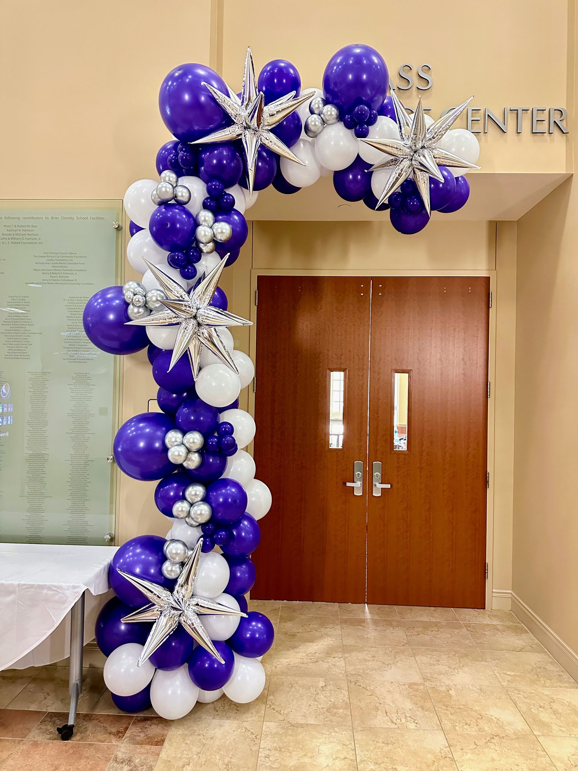 A purple and white balloon arch is hanging over a doorway.