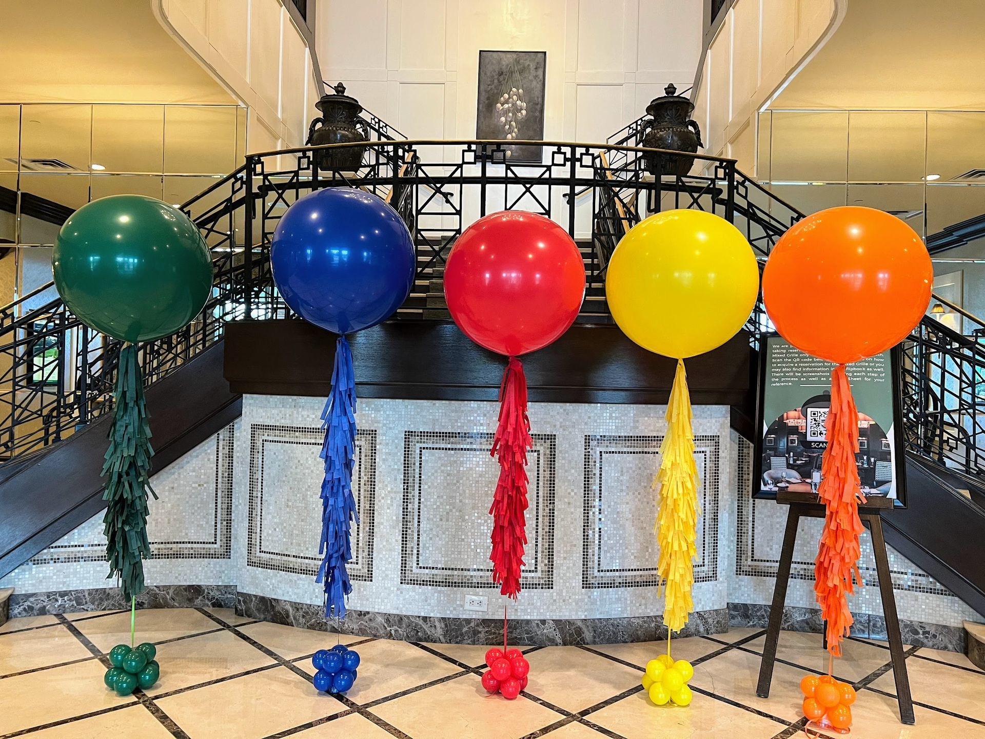 Jumbo colorful helium balloons with fringe tassels decorate a grand hotel lobby staircase. Perfect for all events.