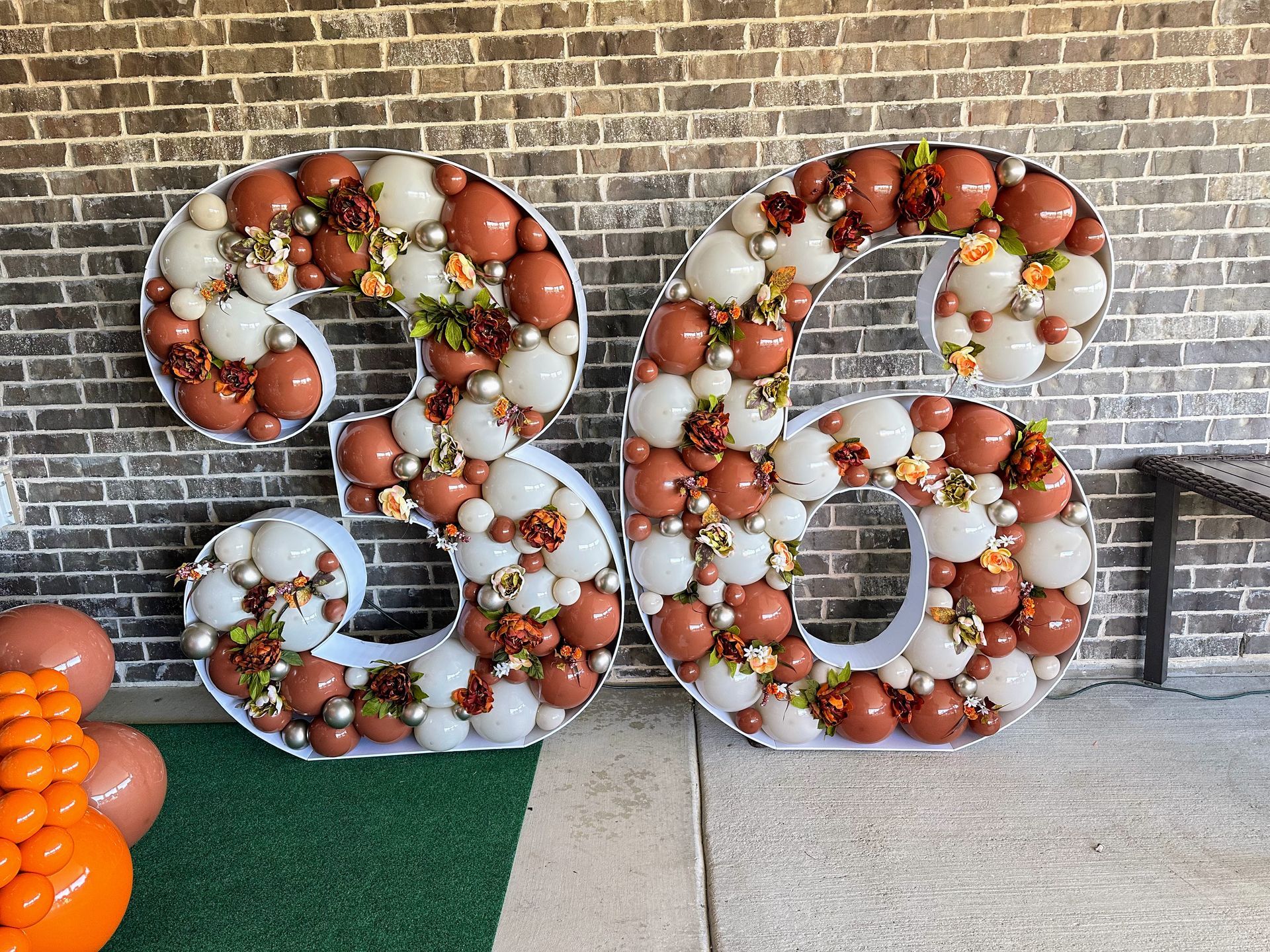 The number 36 is made of balloons and flowers and is sitting in front of a brick wall.
