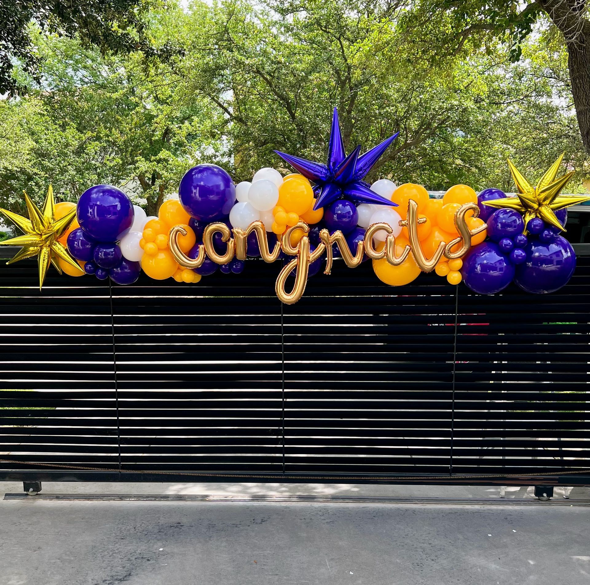 A black gate is decorated with purple and yellow balloons and a congratulations sign.