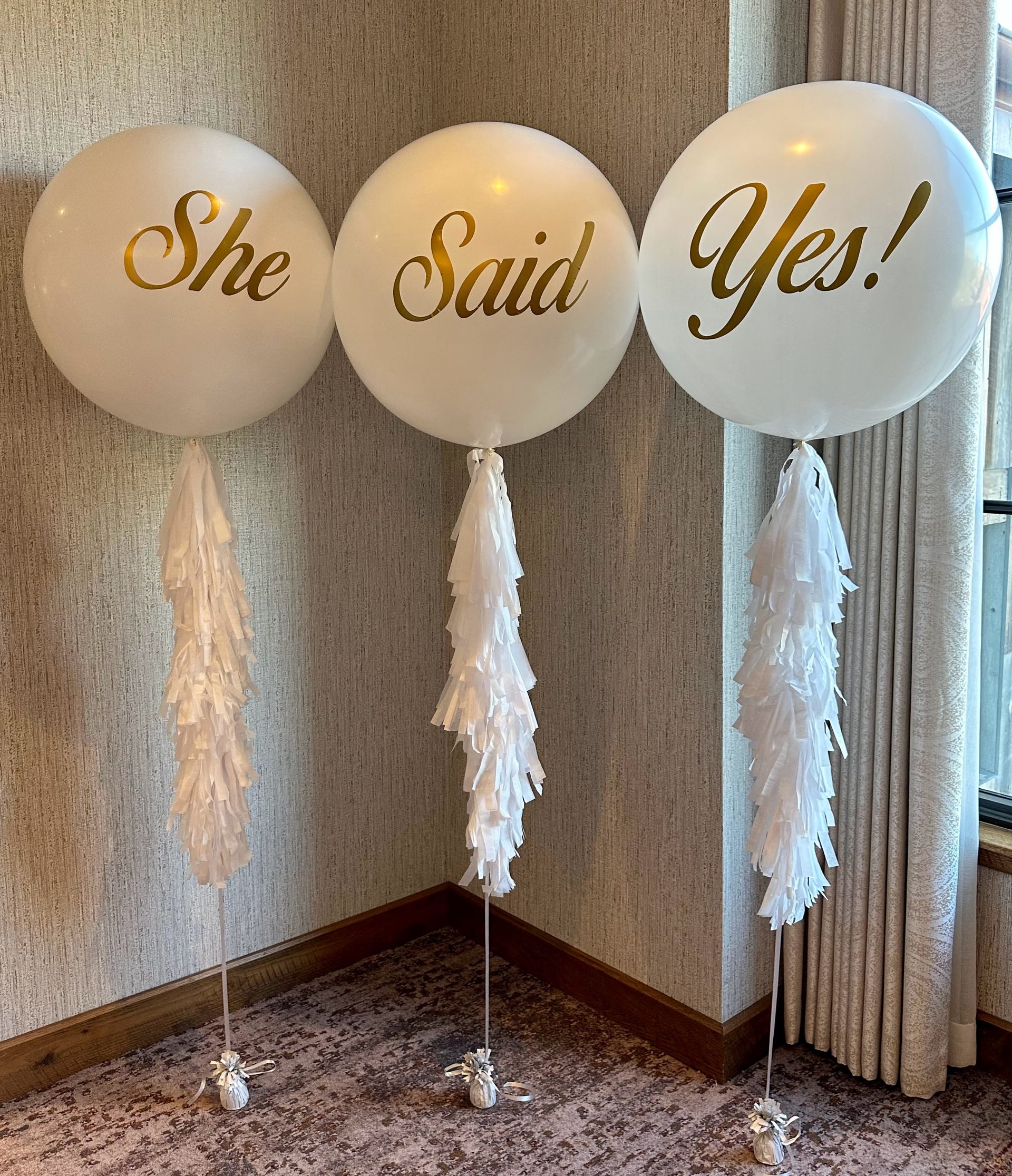Three balloons with the words `` she said yes '' written on them are sitting in a room.