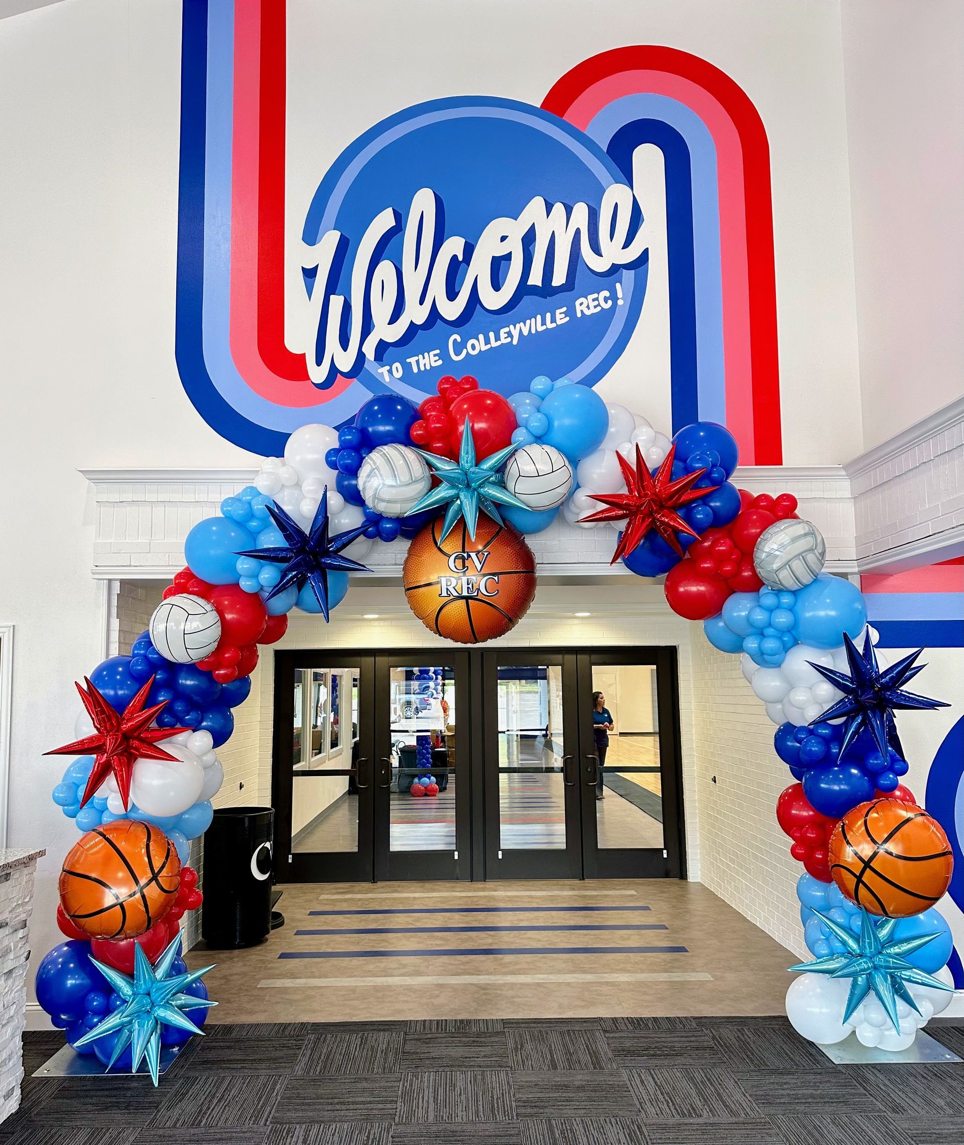 Sports theme balloon arch in red, white and blue balloons with sports accent balloons framing entrance way of gym