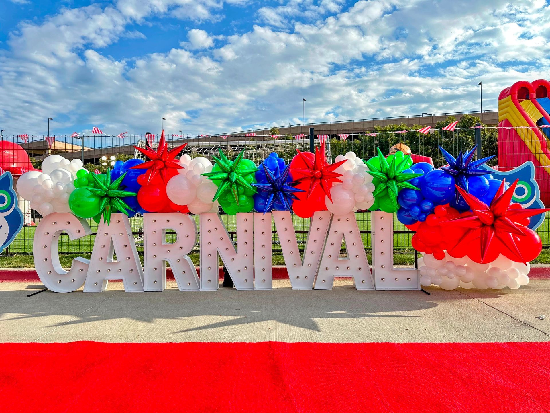 Carnival themed balloon installation featuring large marquee letters and colorful balloon garlands outdoors
