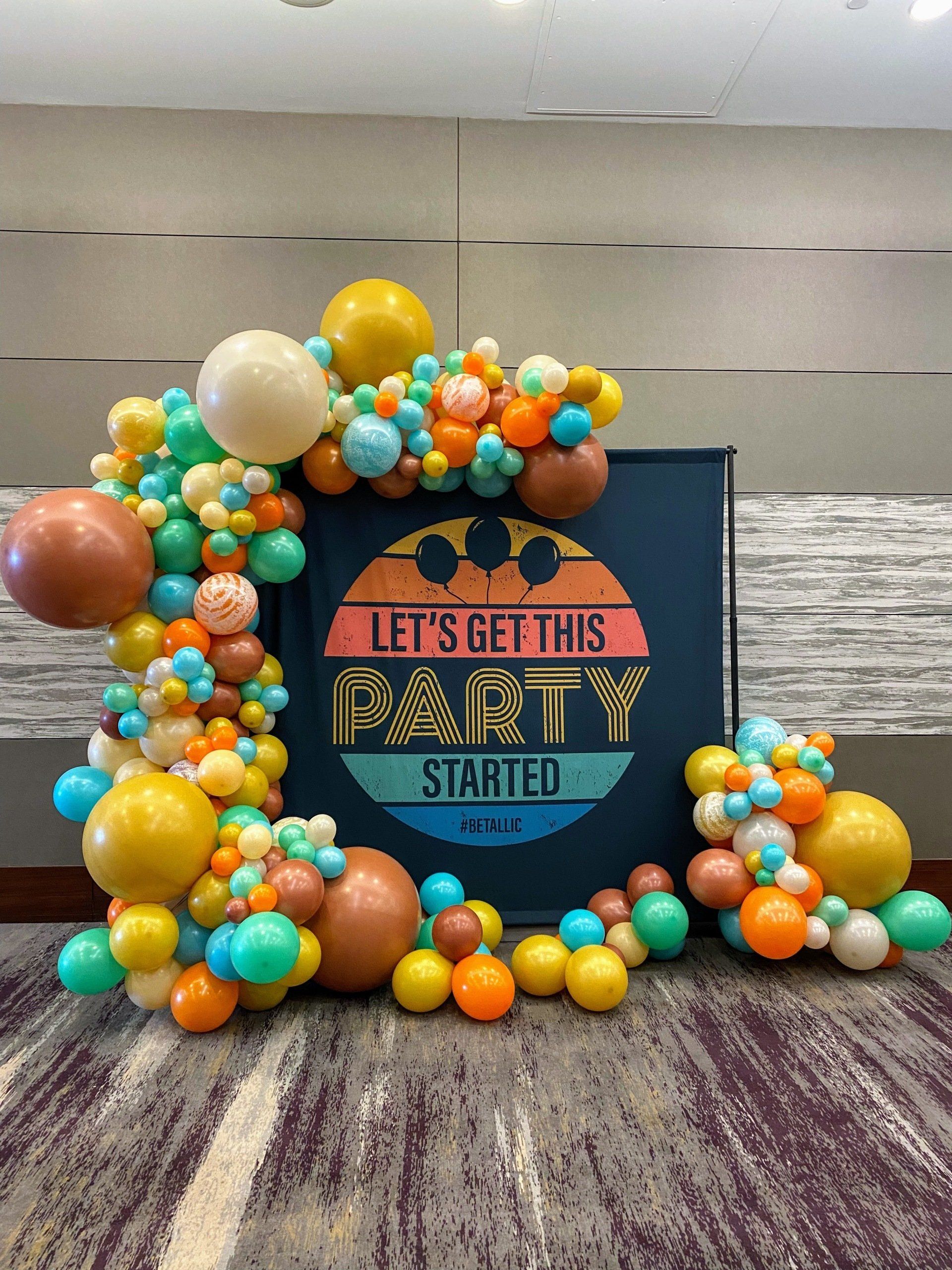 A sign that says let 's get this party started is surrounded by balloons.