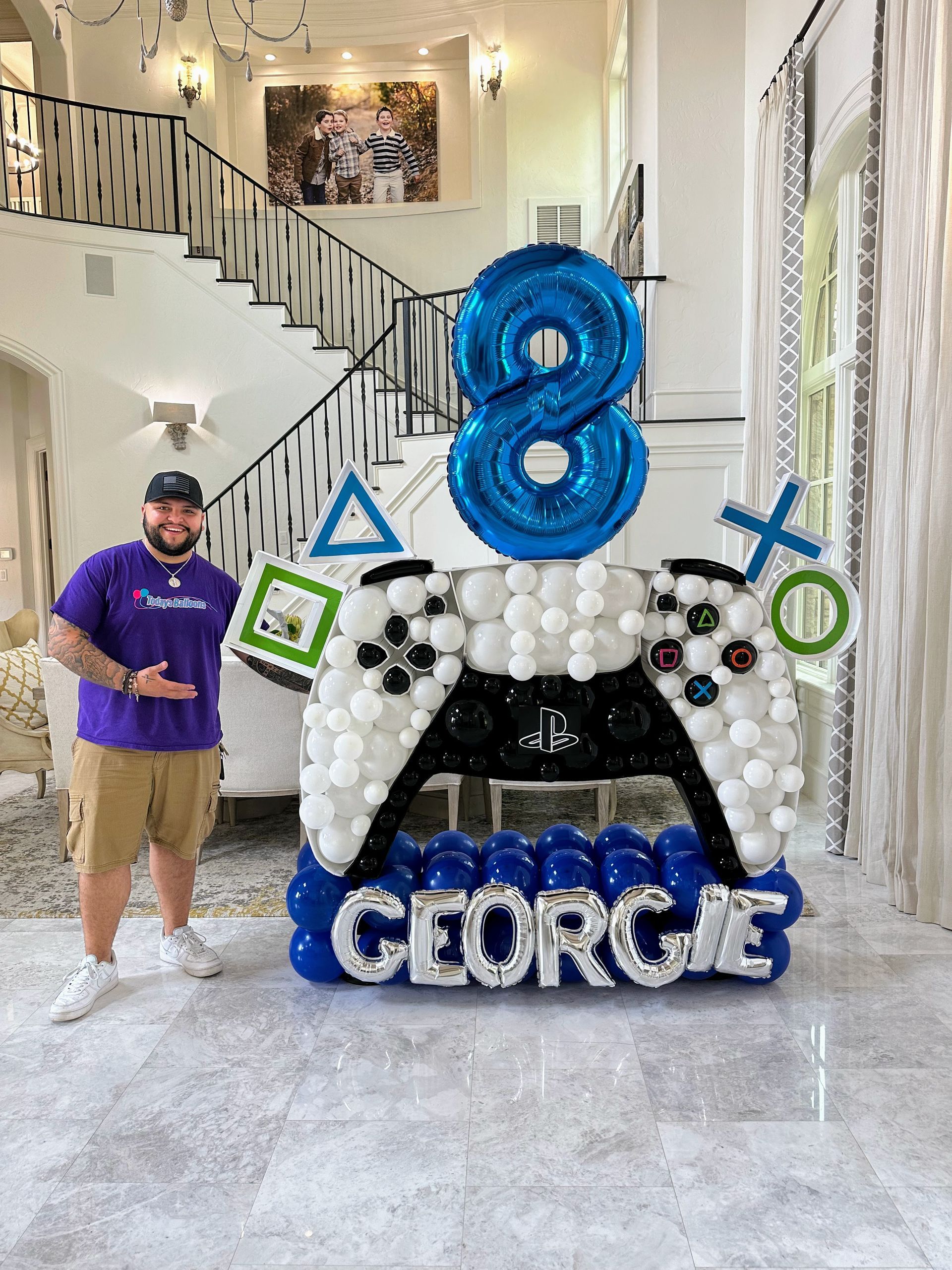 A man is standing in front of a video game controller made of balloons.