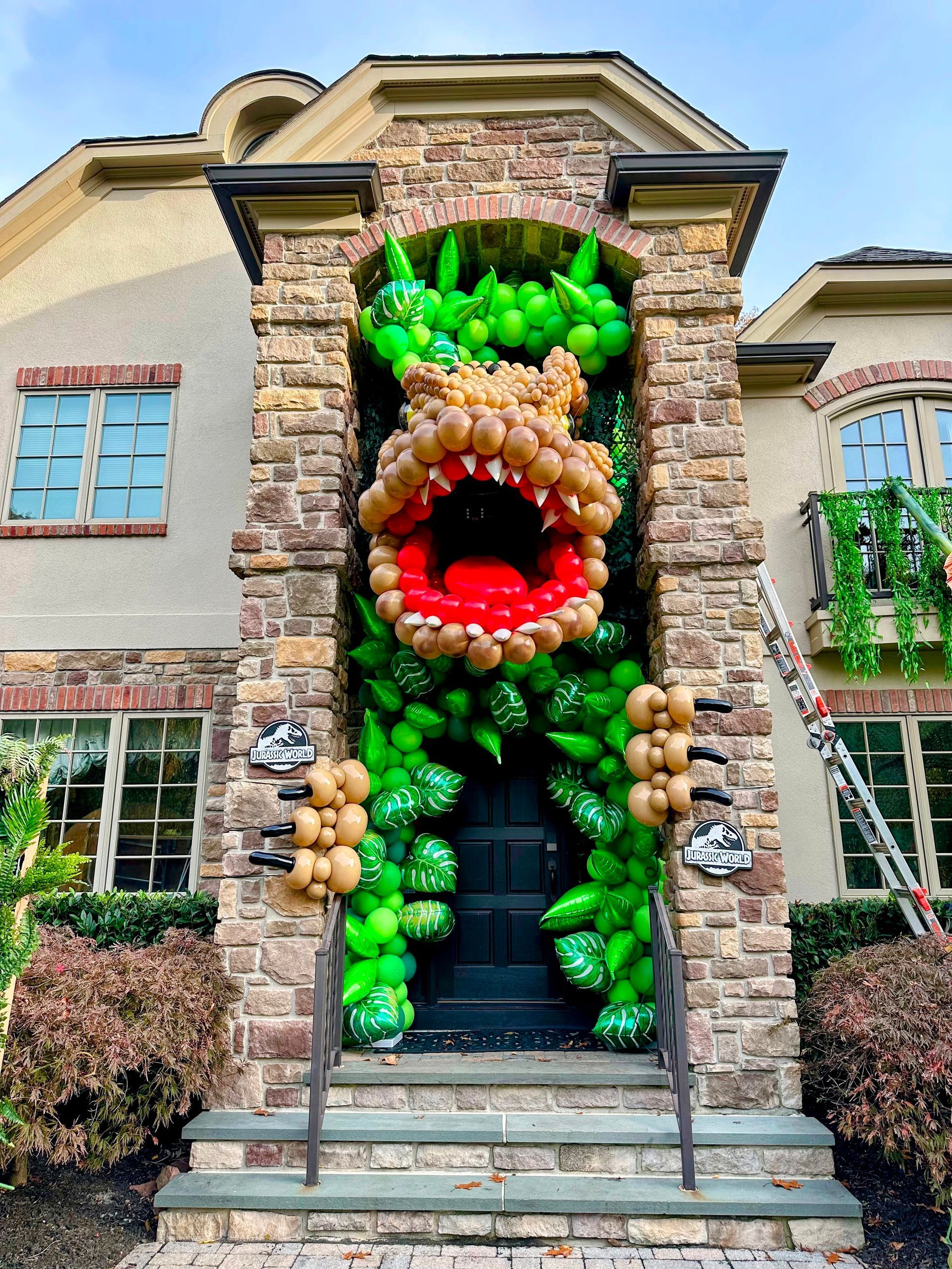 Large balloon sculpture of a light brown T-Rex with green balloon as leaves built into a tall arch way of a two story house
