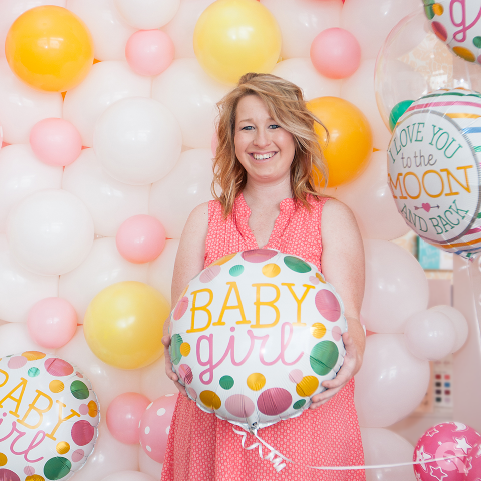 A woman holding a baby girl balloon in front of a wall of balloons