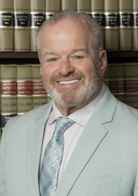 Smiling man in a light gray suit and patterned tie standing before a bookshelf of law books