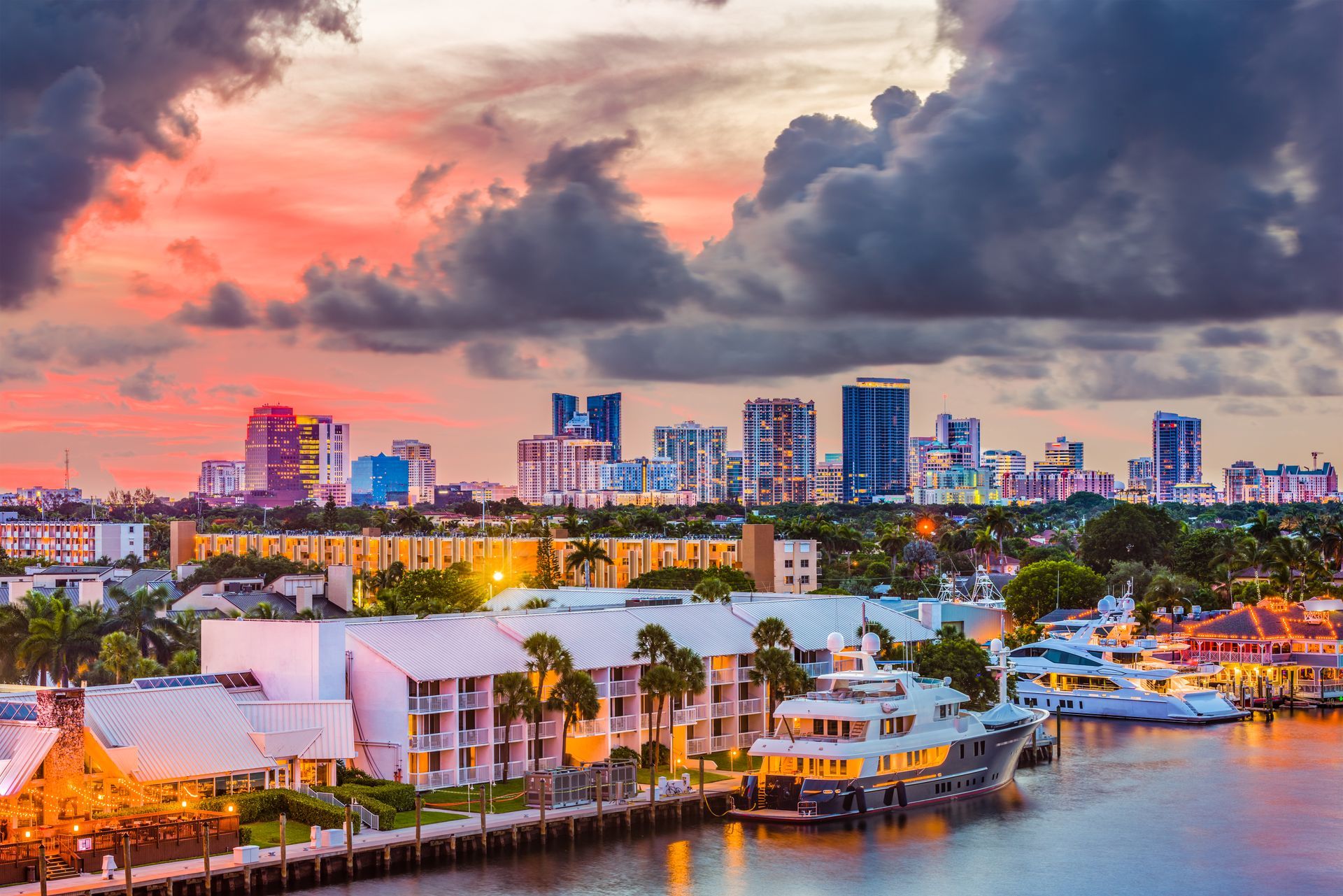 Fort Lauderdale cityscape at sunset with a river, luxury yachts, and tropical vegetation in the foreground.