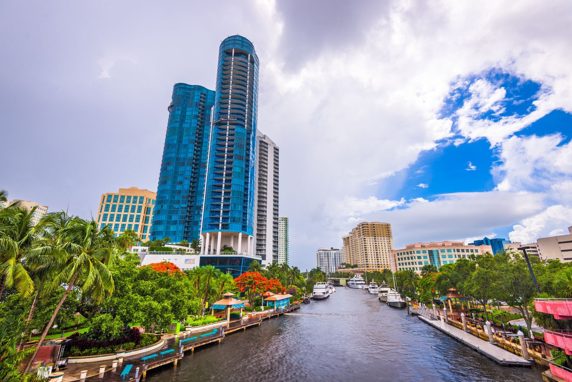 A canal winds between lush trees and blue glass skyscrapers under a partly cloudy sky in a tropical urban setting.
