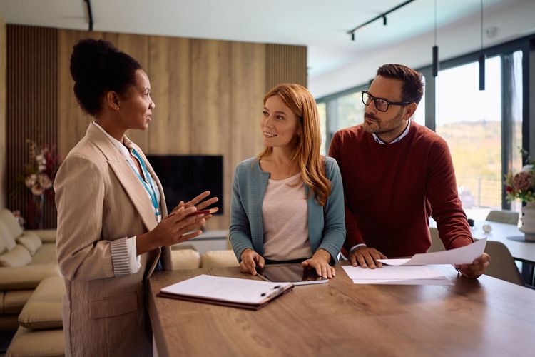Three people discussing documents at a wooden table in a bright, modern living space.