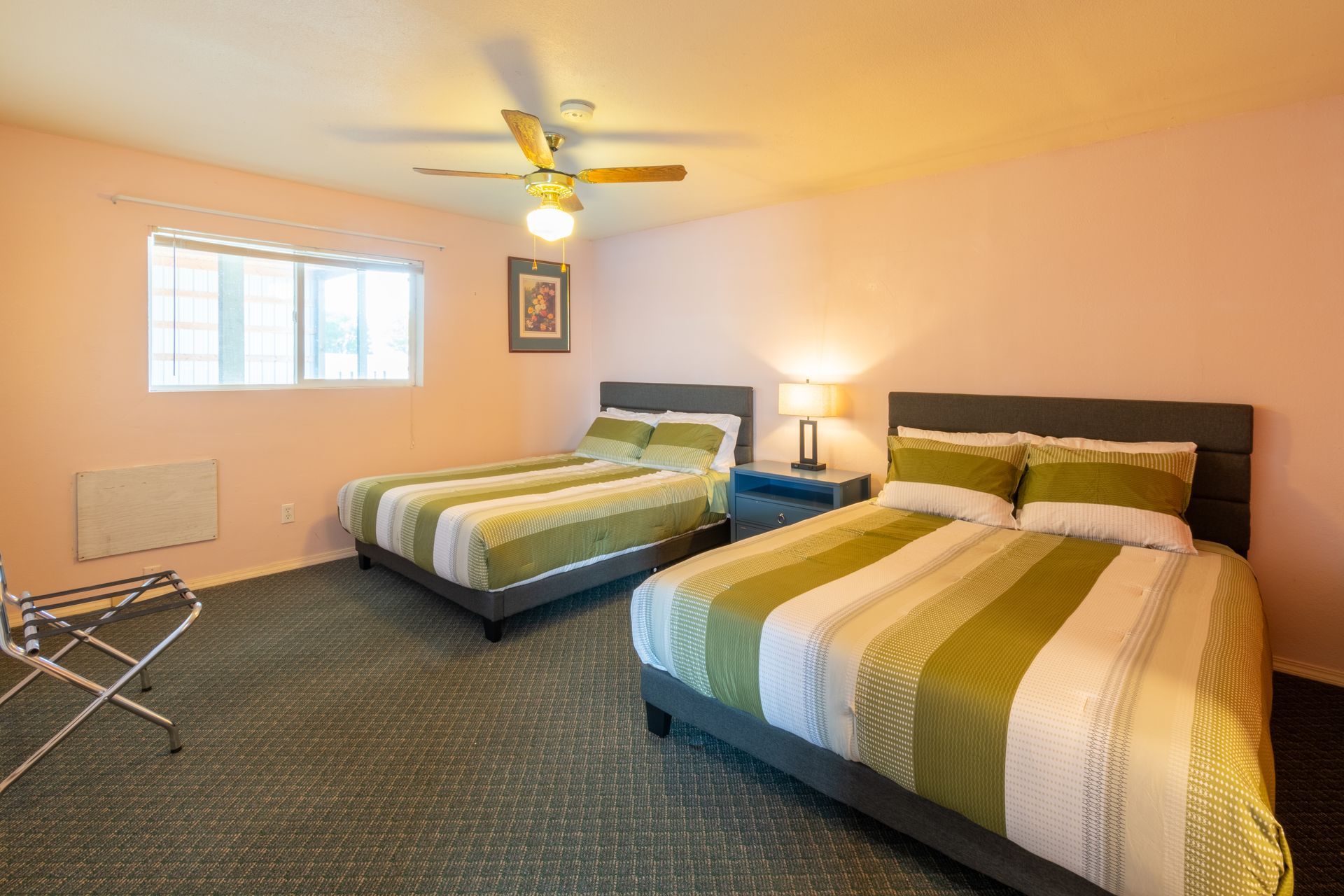 A hotel room with two double beds, featuring green and white striped bedding, set against pink walls and a ceiling fan.