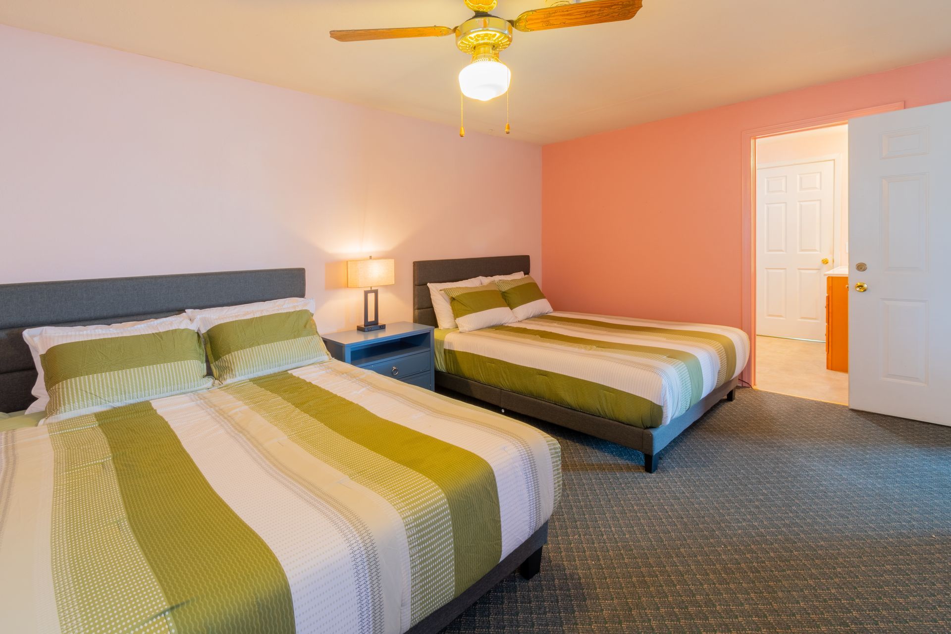 A bedroom with two double beds, pastel pink and coral walls, and a doorway. The beds have green and white striped bedding.