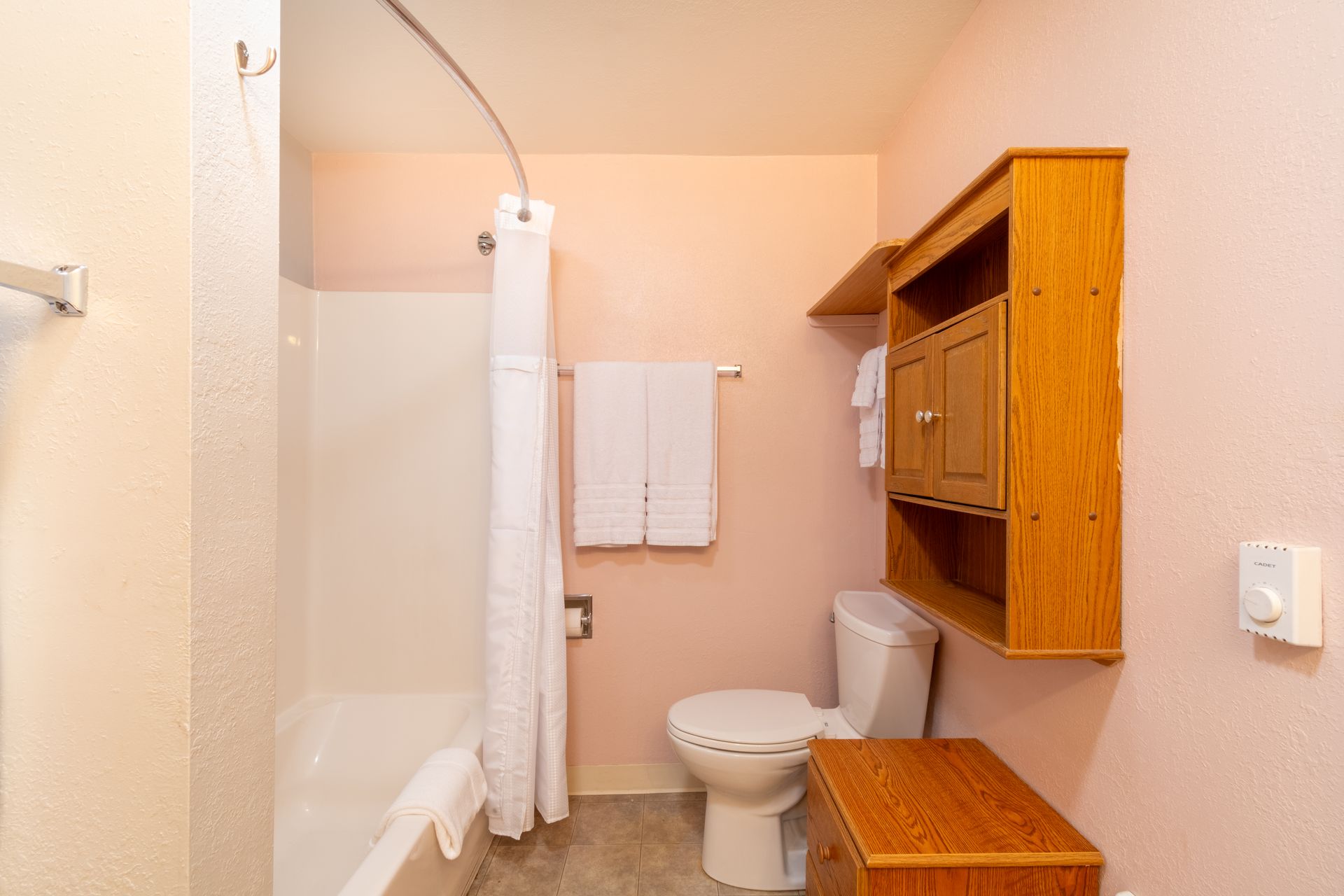 Bathroom with white shower, toilet, and towels.  Wooden cabinet and small dresser stand out against the pink walls.