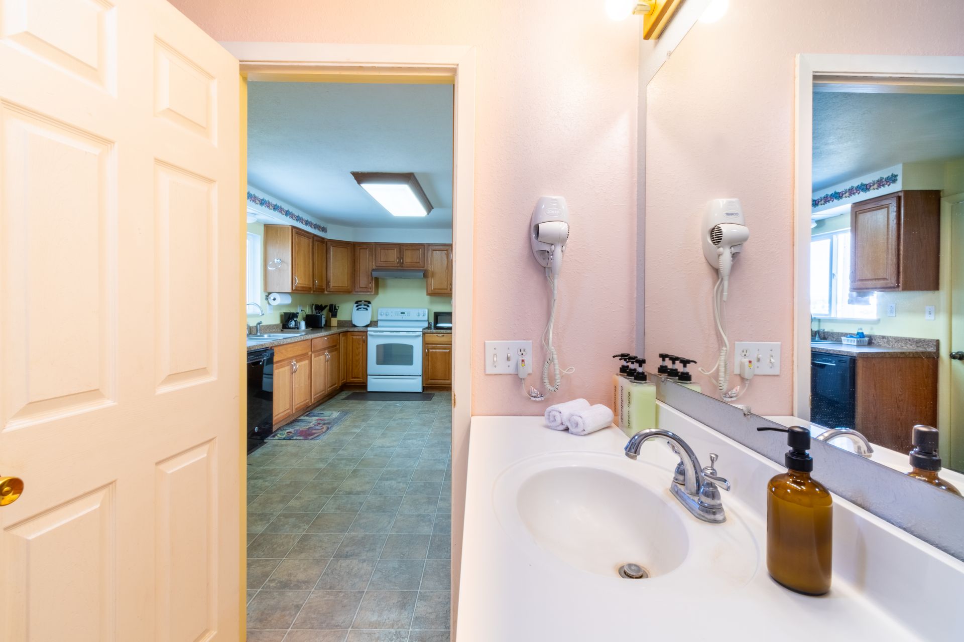 Bathroom with a sink and mirror, reflected kitchen visible in the mirror and through an open doorway. Light pink walls.